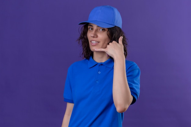 Delivery girl in blue uniform and cap making call me gesture smiling friendly on isolated purple