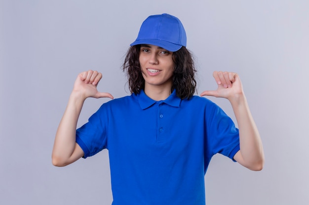 Foto gratuita ragazza delle consegne in uniforme blu e cappuccio che punta a se stessa guardando fiducioso sorridente, soddisfatto di sé e orgoglioso in piedi su bianco