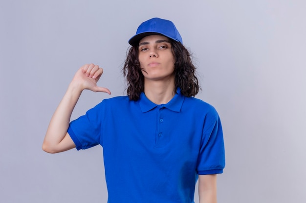 Delivery girl in blue uniform and cap pointing thumb on herself  with serious and confident expression standing over isolated white space