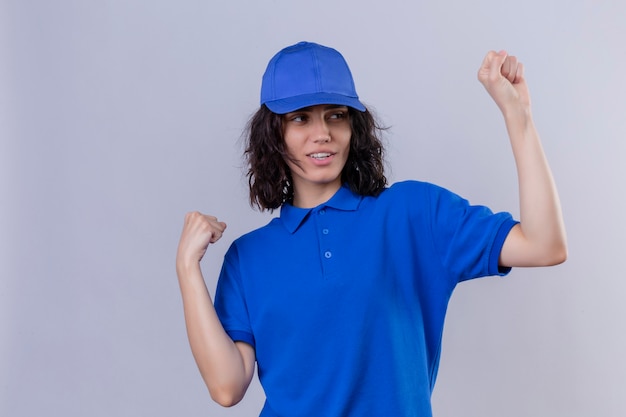 Foto gratuita ragazza delle consegne in uniforme blu e berretto che si rallegra del suo successo e della vittoria stringendo i pugni con gioia felice di raggiungere il suo scopo e obiettivi in piedi su bianco isolato