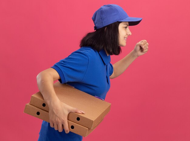 Delivery girl in blue uniform and cap rush running for delivering pizza boxes for customer over pink wall