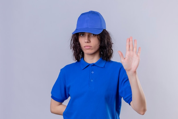 Delivery girl in blue uniform and cap standing with open hand doing stop sign with serious and confident expression, defense gesture on white