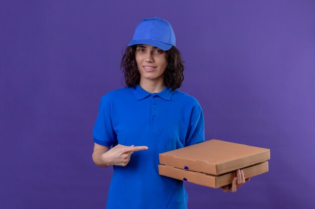 Доставщица в синей форме и кепке стоит с коробками из-под пиццы, указывая на них указательным пальцем, уверенно улыбаясь на изолированном фиолетовом
