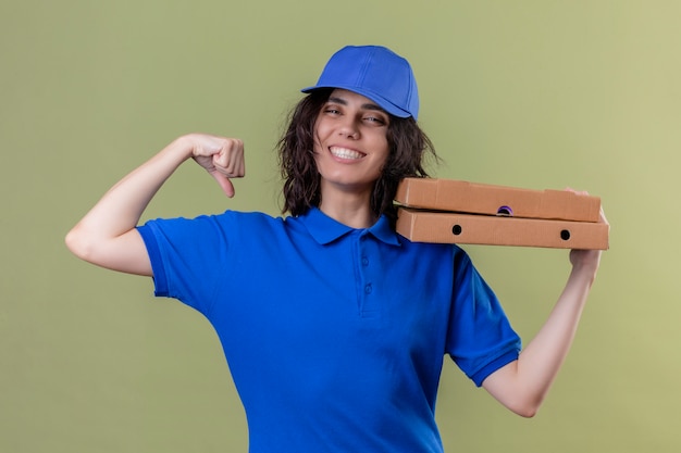 Доставщица в синей форме держит коробки для пиццы, показывая бицепсы, весело улыбаясь, стоя на изолированном зеленом