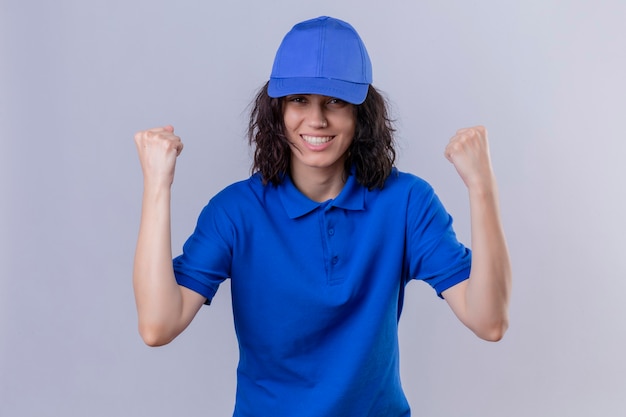Бесплатное фото Доставщица в синей форме и кепке выглядит взволнованной, радуясь своему успеху и победе, сжимая кулаки от радости, счастливая достичь своей цели и целей, стоя на изолированном белом пространстве