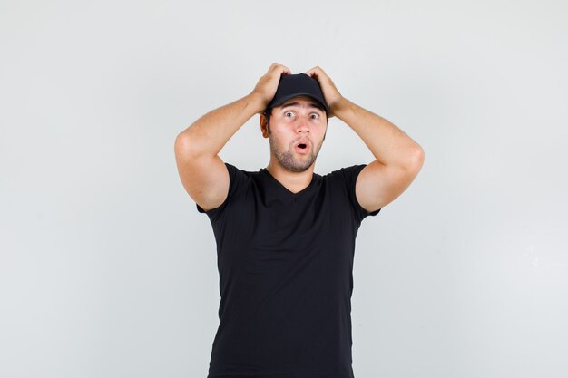 Delivery man in black t-shirt, cap holding hands on head and looking shocked