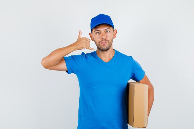 Delivery man in blue t-shirt, cap holding cardboard box with phone gesture