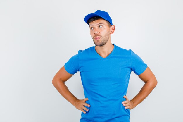 Delivery man in blue t-shirt, cap looking aside with hands on waist and looking pensive