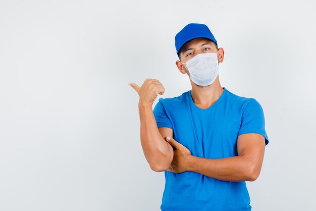 Delivery man in blue t-shirt, cap, mask pointing thumb to side and looking confident