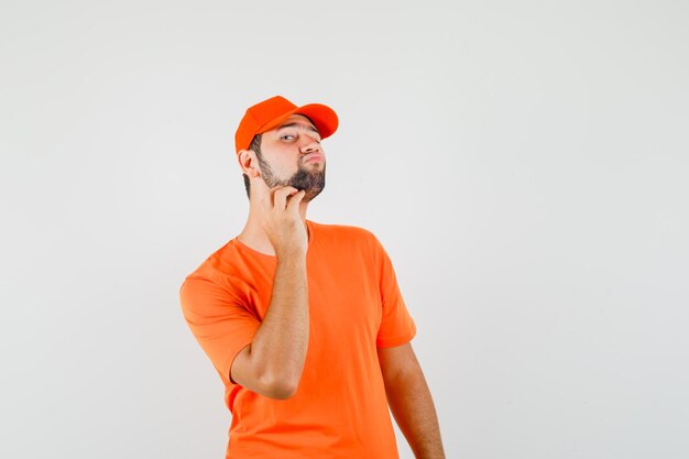 Доставщик проверяет кожу лица, касаясь его бороды в оранжевой футболке, кепке и выглядит стильно, вид спереди.
