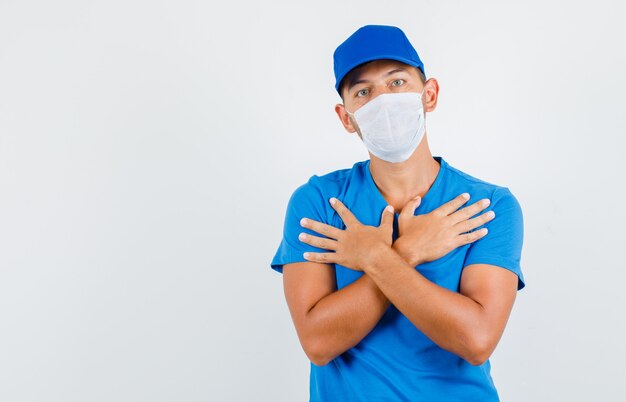 Delivery man crossing arms on chest in blue t-shirt, cap, mask and looking helples