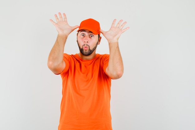 Delivery man doing funny gesture with hands as ears in orange t-shirt, cap front view.