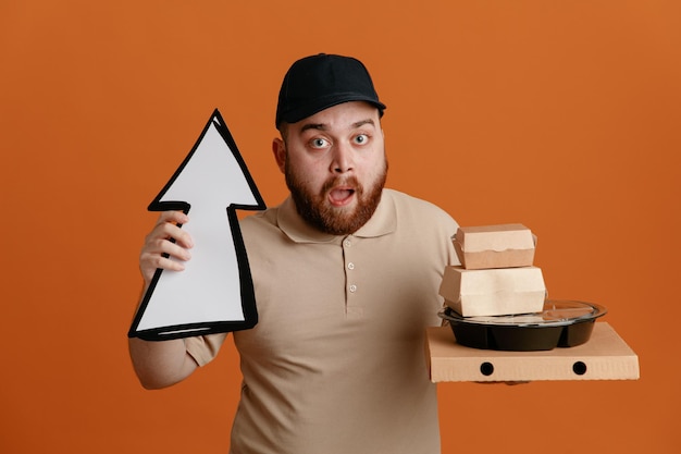 Free photo delivery man employee in black cap and blank tshirt uniform holding arrow and food containers looking at camera amazed and surprised standing over orange background