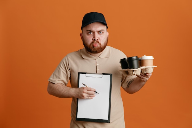 Dipendente uomo di consegna in berretto nero e maglietta vuota uniforme che tiene tazze di caffè e appunti con penna in attesa di firma accigliata guardando la fotocamera in piedi su sfondo arancione