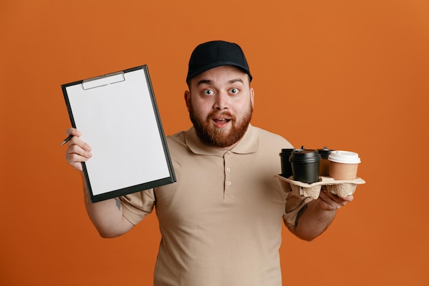 Dipendente dell'uomo delle consegne in berretto nero e uniforme della maglietta vuota che tiene tazze di caffè e appunti con la penna in attesa di una firma guardando la fotocamera felice e sorpreso in piedi su sfondo arancione