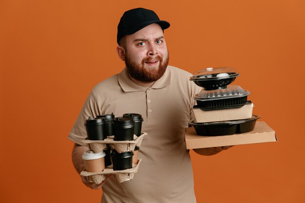 Delivery man employee in black cap and blank tshirt uniform holding coffee cups and food containers looking at camera annoyed and tired standing over orange background