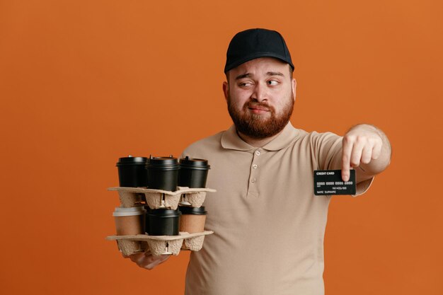 Сотрудник службы доставки в черной кепке и пустой футболке держит кофейные чашки, показывая кредитную карту, смотрит в сторону со скептическим выражением лица, стоя на оранжевом фоне