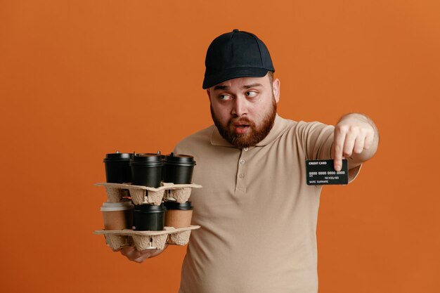 Сотрудник доставщика в черной кепке и чистой форме футболки держит кофейные чашки, показывая кредитную карту, выглядит смущенным, стоя на оранжевом фоне
