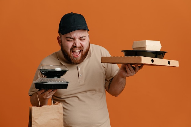 オレンジ色の背景の上に立って怒って狂った狂牛病の叫びと叫びと叫んで紙袋と食品容器を保持している黒い帽子と空白のTシャツの制服を着た配達人の従業員