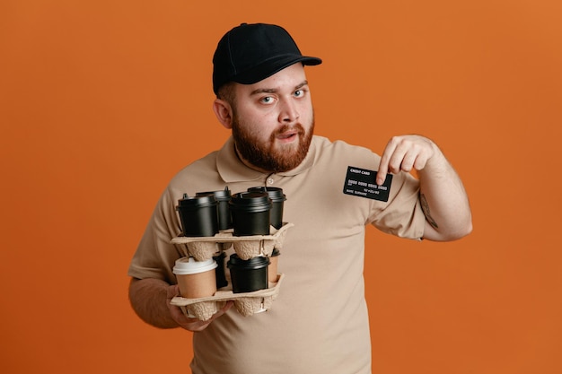 Сотрудник службы доставки в черной кепке и чистой форме футболки держит кофейные чашки, показывая кредитную карту, смотрит в камеру с уверенным выражением лица, стоя на оранжевом фоне