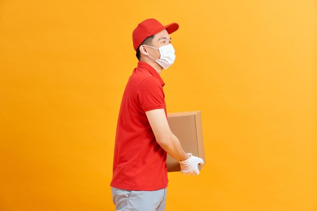 赤い帽子の空白のtシャツの制服のフェイスマスクの手袋の配達人の従業員は、黄色の背景で隔離の段ボール箱を保持します。