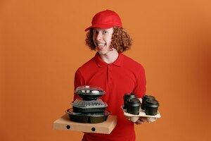 Сотрудник службы доставки в красной кепке, пустой футболке, держит коробку с пиццей, контейнеры с едой и кофейные чашки, смотрит в камеру, счастливый и радостный, высунув язык, стоя на оранжевом фоне