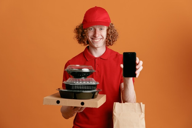 Бесплатное фото Сотрудник доставщика в красной кепке, пустой футболке, держит коробку для пиццы, контейнеры для еды с бумажным пакетом, показывая смартфон, счастливый и позитивный, весело улыбающийся, стоя на оранжевом фоне