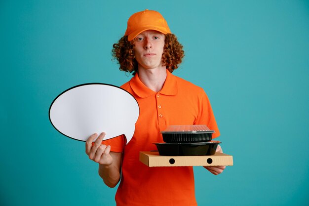 Delivery man employee in orange cap blank tshirt uniform holding empty speech bubble and food containers looking at camera with confident expression standing over blue background