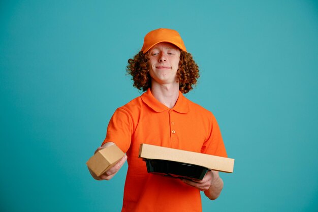 Delivery man employee in orange cap blank tshirt uniform holding pizza box and food containers smiling confident standing over blue background