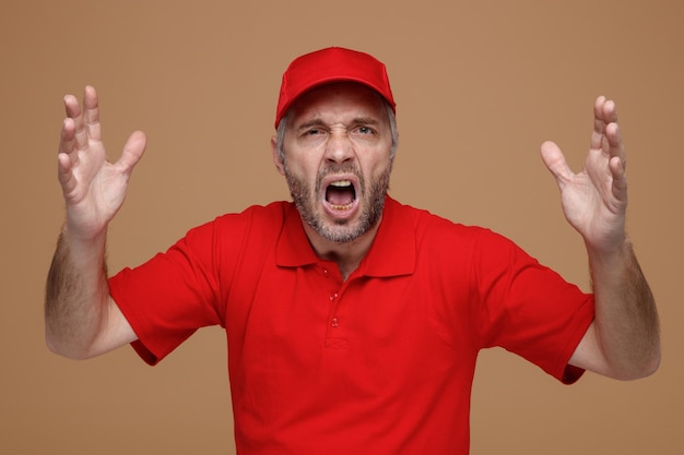 赤い帽子の空白のTシャツの制服を着た配達人の従業員は茶色の背景の上に立っている腕を上げて怒って欲求不満の叫び