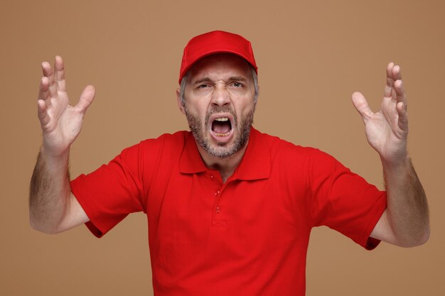 Delivery man employee in red cap blank tshirt uniform angry and frustrated yelling with raised arms standing over brown background