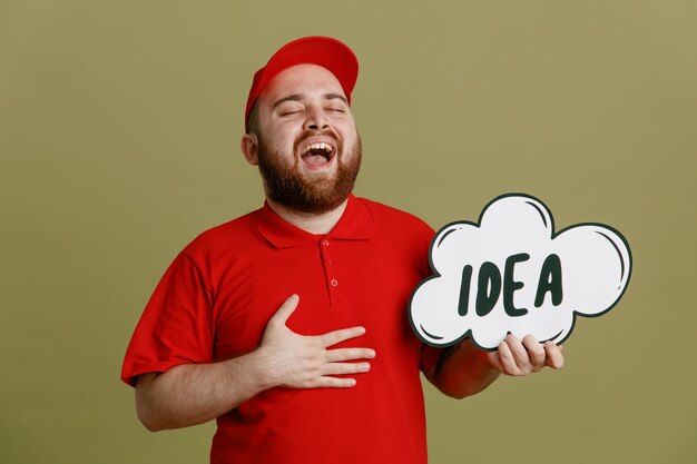 Delivery man employee in red cap blank tshirt uniform holding bubble speech with word idea laughing out happy and excited standing over green background