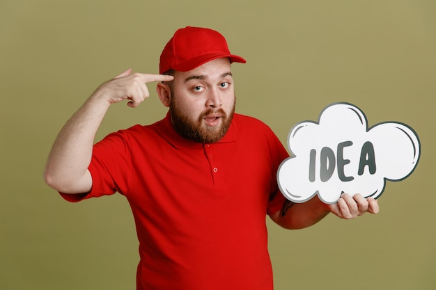 Delivery man employee in red cap blank tshirt uniform holding bubble speech with word idea looking at camera pointing with index finger at his head standing over green background