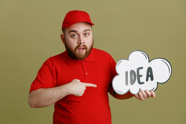 Delivery man employee in red cap blank tshirt uniform holding bubble speech with word idea pointing with index finger at it looking surprised standing over green background