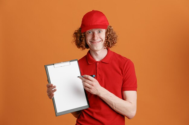 Delivery man employee in red cap blank tshirt uniform holding clipboard with pen looking at camera happy and joyful smiling cheerfully waiting for signature standing over orange background