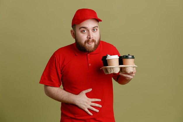 Delivery man employee in red cap blank tshirt uniform holding coffee cups looking at camera happy and joyful sticking out tongue standing over green background