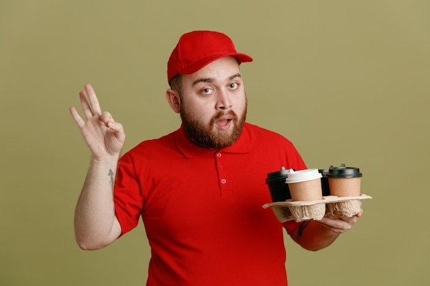 Delivery man employee in red cap blank tshirt uniform holding coffee cups looking at camera happy and positive doing ok sign standing over green background