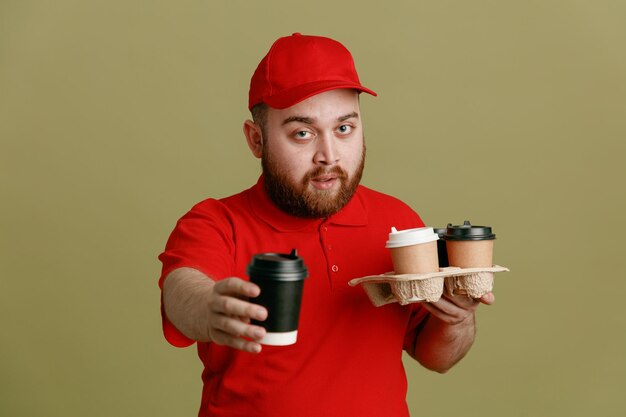 Сотрудник доставщика в красной кепке с пустой футболкой держит чашки кофе, предлагая чашку, протягивая руку, выглядя уверенно, стоя на зеленом фоне