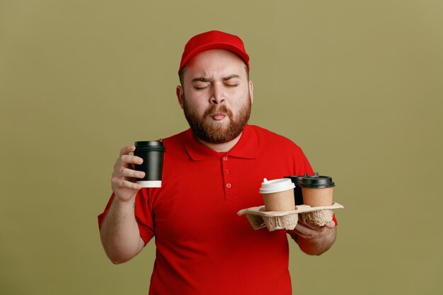 赤い帽子の空白のTシャツの制服を着た配達人の従業員は、緑の背景の上に立って目を閉じて幸せで満足してコーヒーを味わうコーヒーカップを保持しています