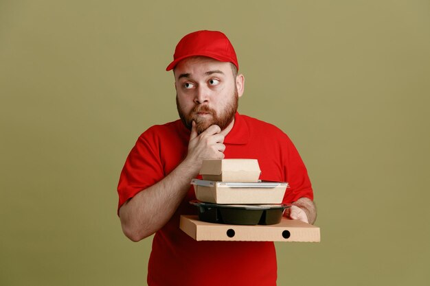 緑の背景の上に立っている彼のあごに手で考えて物思いにふける表情で脇を見て食品容器とピザボックスを保持している赤い帽子の空白のTシャツの制服を着た配達人の従業員