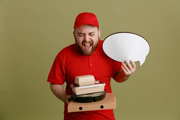 Delivery man employee in red cap blank tshirt uniform holding food containers and pizza box with empty speech bubble happy and excited standing over green background
