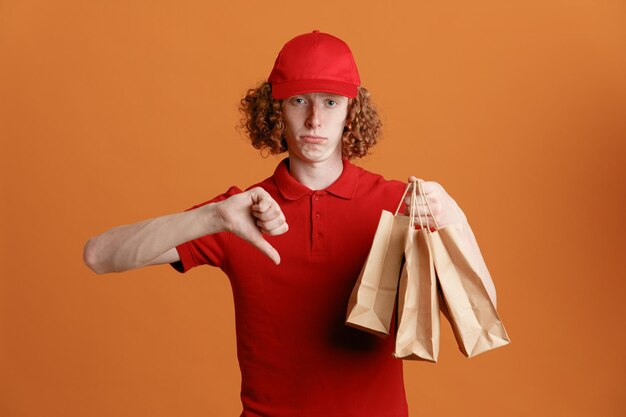 Сотрудник доставщика в красной кепке с пустой футболкой держит бумажные пакеты, недовольно смотрит в камеру, показывая большой палец вниз, недовольный стоя на оранжевом фоне