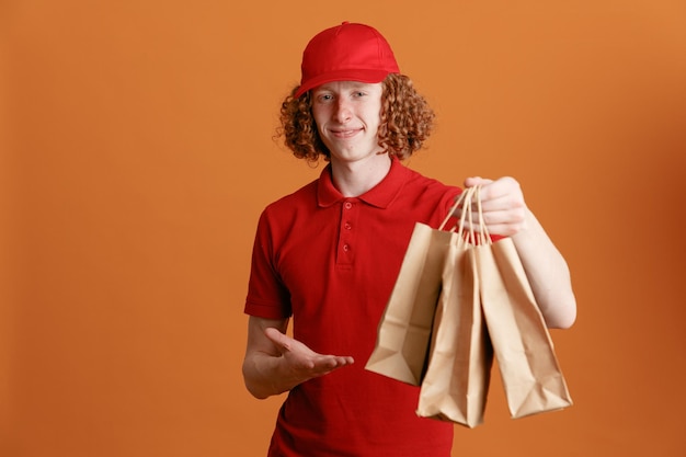 Uomo di consegna dipendente in berretto rosso maglietta vuota uniforme in possesso di sacchetti di carta presentando con il braccio guardando la fotocamera felice e positivo sorridente fiducioso in piedi su sfondo arancione