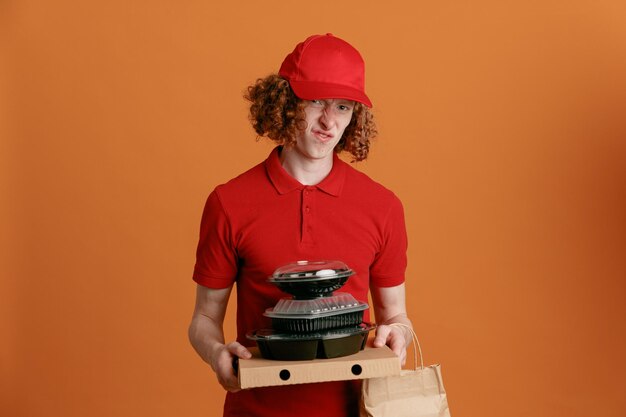 빨간 모자를 쓴 빈 티셔츠를 입은 배달원 직원이 피자 상자 음식 용기를 들고 종이 봉지를 들고 오렌지색 배경 위에 서서 실망한 카메라를 바라보고 있다
