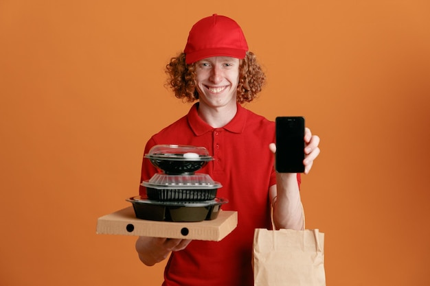 Foto gratuita fattorino dipendente in berretto rosso maglietta vuota uniforme che tiene scatola della pizza contenitori per alimenti con sacchetto di carta che mostra smartphone felice e positivo sorridente allegramente in piedi su sfondo arancione