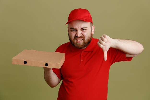 빨간 모자를 쓴 빈 티셔츠를 입은 배달원 직원이 피자 상자를 들고 카메라를 쳐다보며 녹색 배경 위에 엄지손가락을 아래로 내리고 있는 짜증난 표정에 불만을 품고 있다