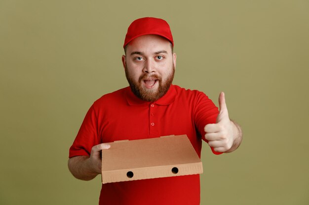 Delivery man employee in red cap blank tshirt uniform holding pizza box looking at camera happy and positive showing thumb up standing over green background