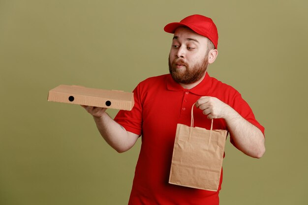 Сотрудник доставщика в красной кепке с пустой футболкой держит коробку для пиццы и бумажный пакет, глядя на коробку, смущенный и удивленный, стоя на зеленом фоне