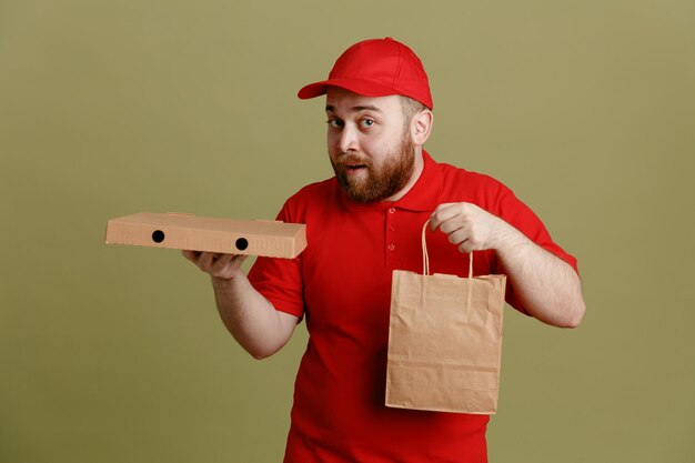 緑の背景の上に立って幸せで驚いたカメラを見てピザボックスと紙袋を保持している赤い帽子の空白のTシャツの制服を着た配達人の従業員