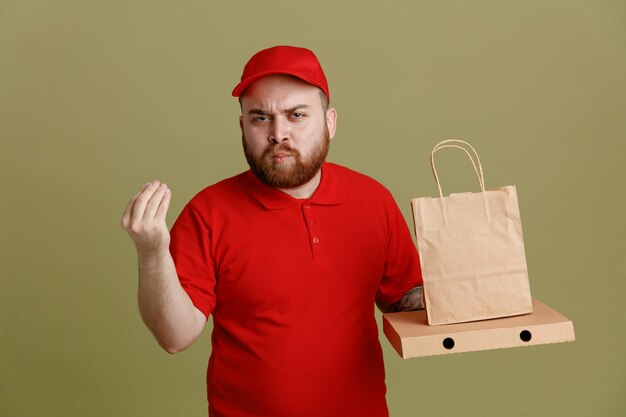 빨간 모자 빈 티셔츠 유니폼을 입은 배달원 직원이 피자 상자와 종이 가방을 들고 찡그린 얼굴로 카메라를 쳐다보며 돈을 벌기 위해 손가락을 문지르는 제스처로 지불을 기다리고 있다
